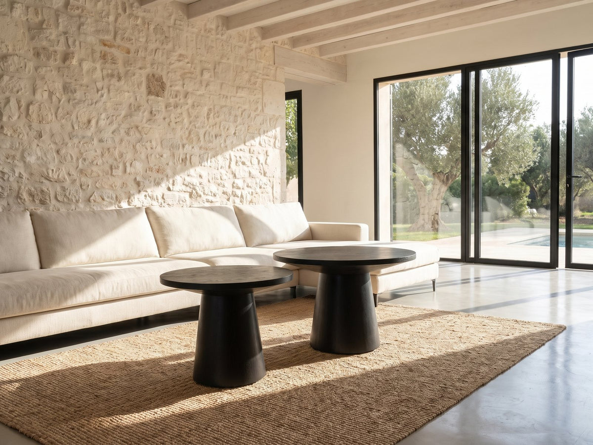 Acqui 29-Inch Round Ebony Oak Veneer Pedestal Coffee and End Table Set seen from the side, on a jute rug in front of a linen sofa in a poolside villa