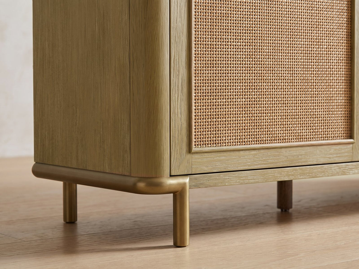 Close-up of the Alassio 80-Inch Sand Wash Ash and Rattan Media Console: the rounded end of the case on its aged-brass sled leg