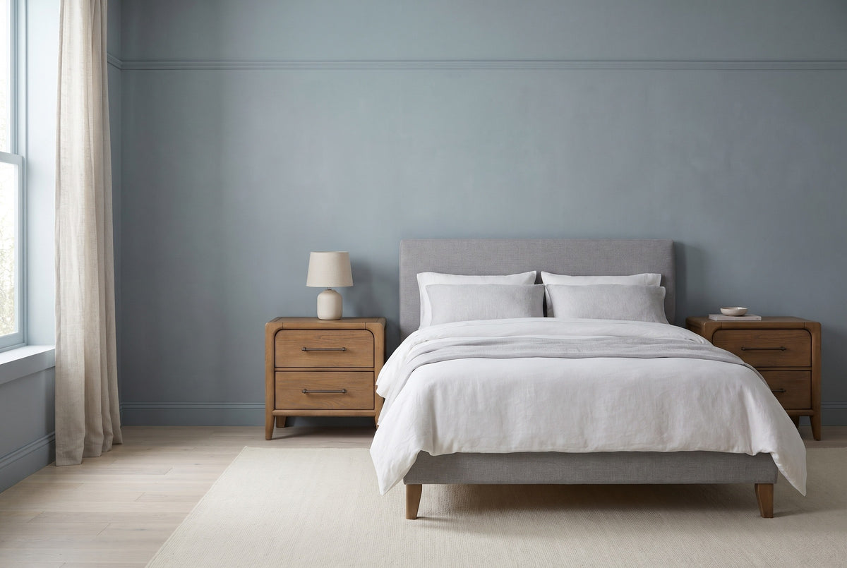 The Alghero set of two brown oak nightstands, one on each side of a bed with a light grey headboard in a Brooklyn bedroom with a dusty blue-grey wall and a tall window