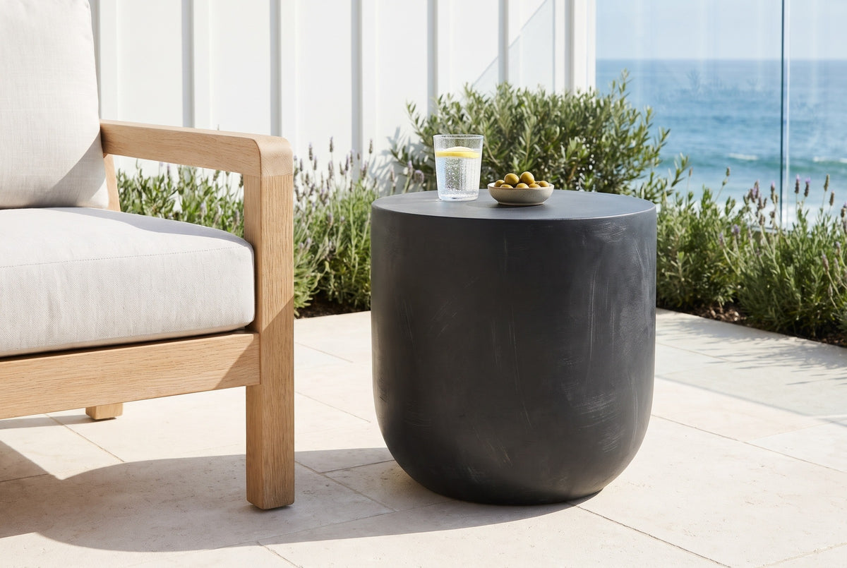 Altavilla matte black side table on limestone pavers on a Laguna Beach terrace beside an oak lounge chair, the Pacific behind (chair, glass and olives not included)