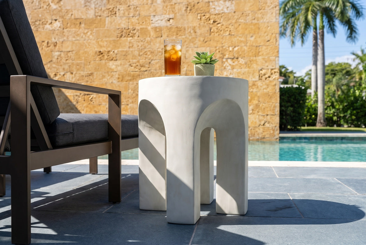 Aragona antique white arched side table with iced tea and a succulent beside a bronze lounge chair on a bluestone pool terrace in Coral Gables, a coral stone wall behind (chair and decor not included)