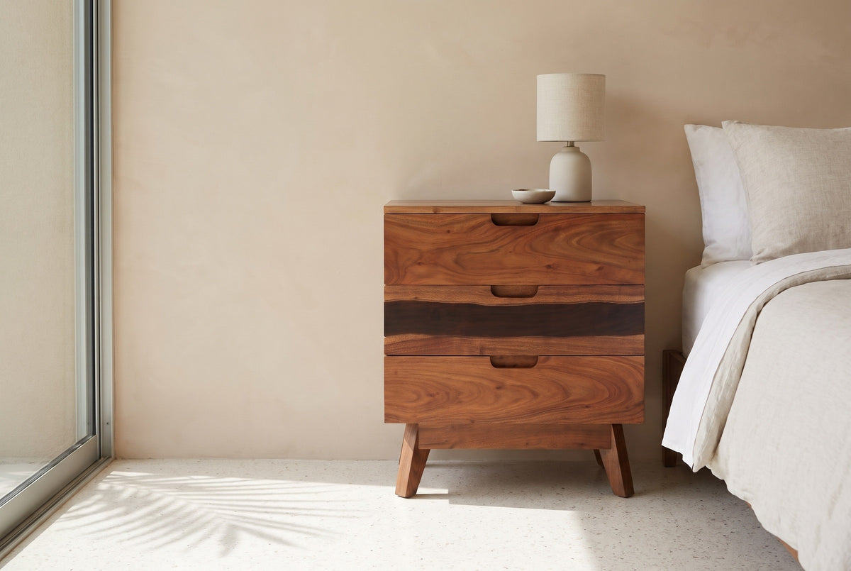 Arborea acacia three-drawer nightstand with a ceramic lamp beside a bed in a Palm Springs bedroom with a sand-beige wall and a tall glass window