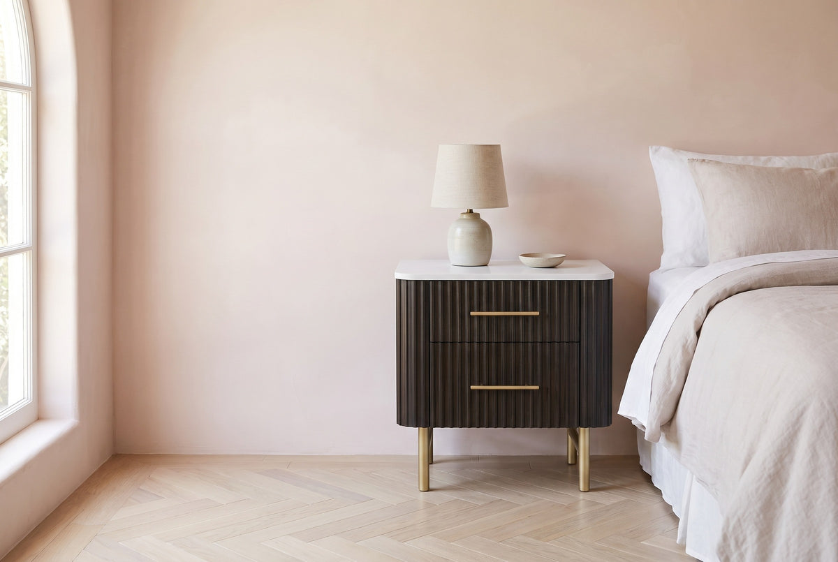 Aritzo espresso reeded nightstand with a white stone top, gold legs and a ceramic lamp beside a bed in a Los Angeles bedroom with a blush-pink plaster wall and an arched window