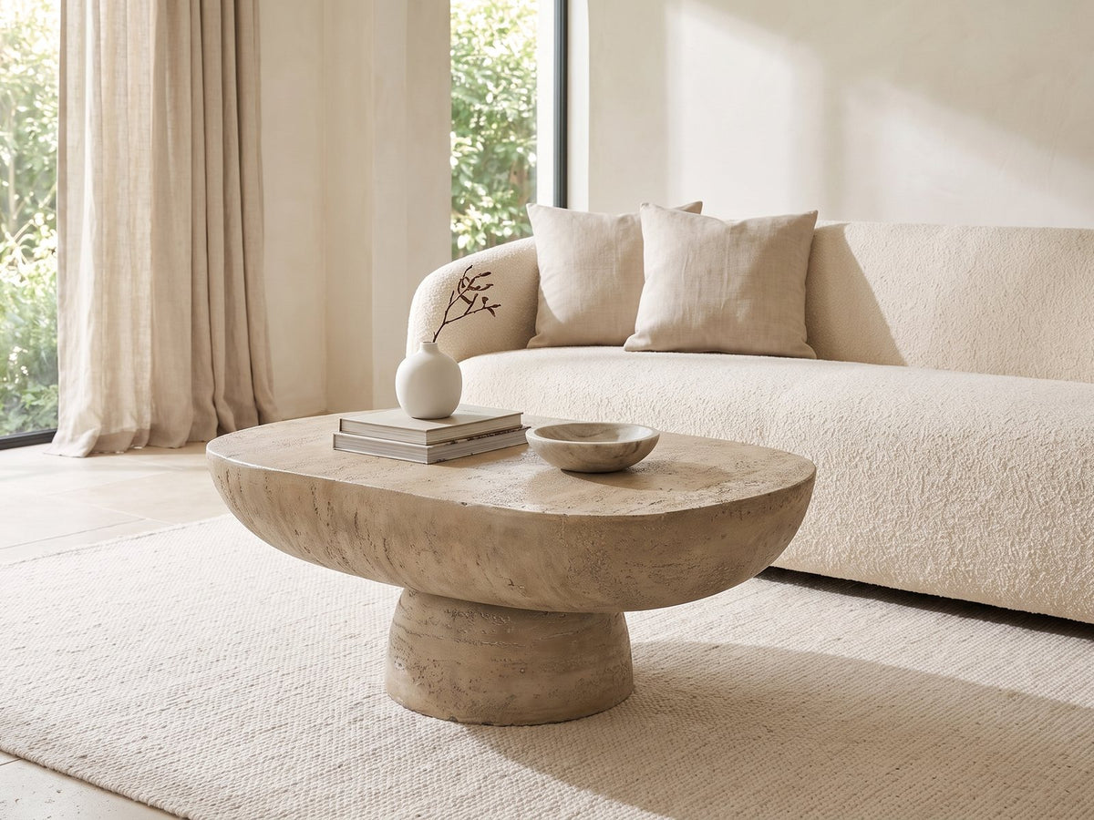 Asiago Faux Travertine Oval Mushroom Coffee Table styled with books, a vase and a stone bowl in front of a cream sofa in a bright plaster room