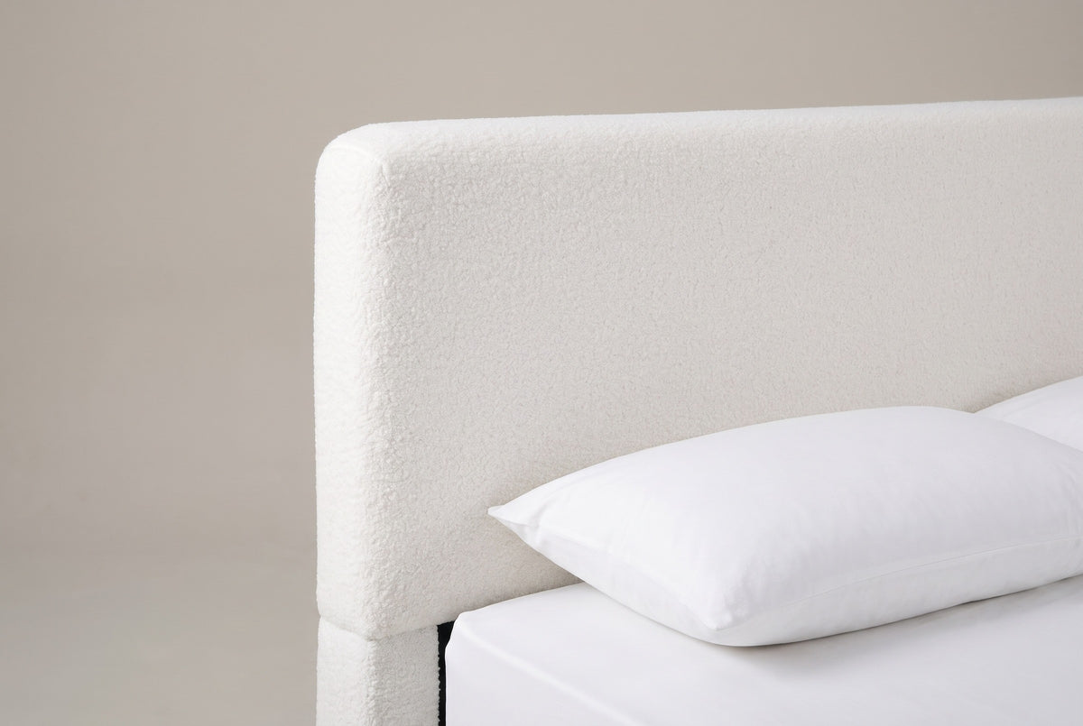 One top corner of the Atessa headboard: a flat padded panel in white boucle with a softly rounded corner above a white pillow