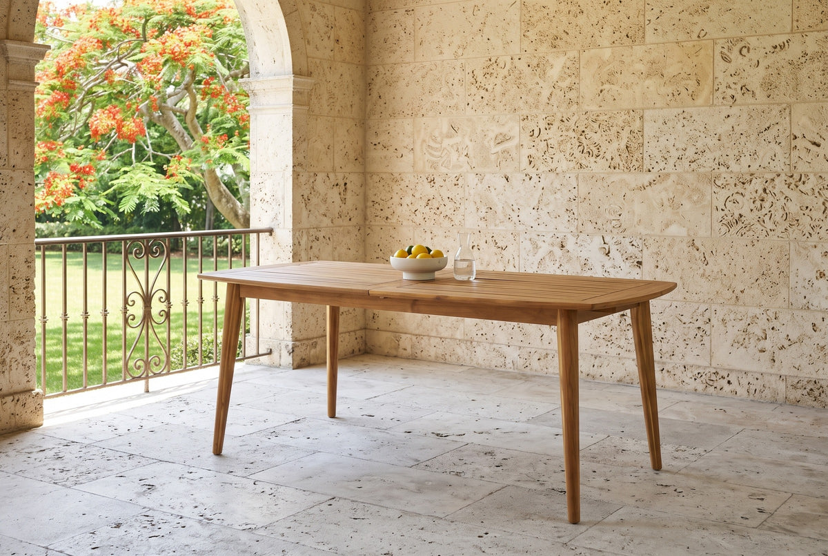 Augusta acacia dining table under a stone loggia with a coral-stone wall and pale limestone floor, a lawn and a flowering tree through the arch (chairs not included, fruit bowl and carafe not included)