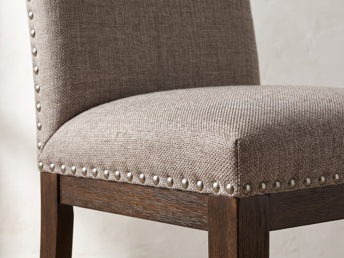 Close-up of the Bagnara 76.5 to 94.5-Inch Rustic Oak Double Pedestal Dining Set with 4 Chairs: the taupe linen-look seat, pewter nailheads and dark brown seat rail of a chair