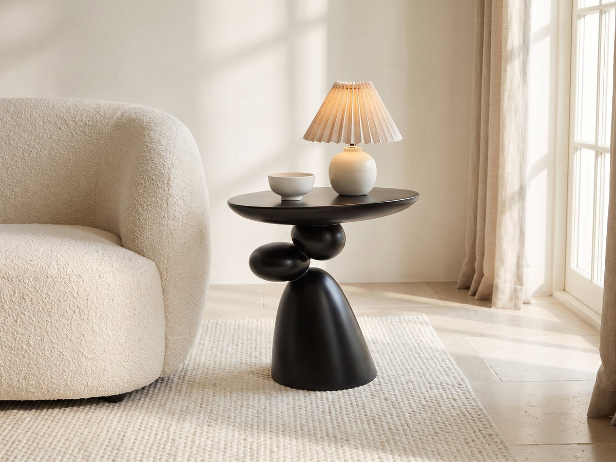 Bitetto Black Fiberglass Stacked Pebble Side Table with a lamp and a ceramic bowl beside a cream boucle armchair in a bright plaster room