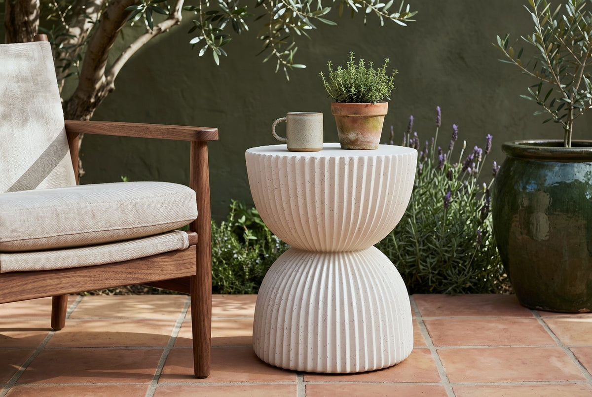 Bivona beige ribbed hourglass side table with a stoneware mug and a pot of thyme beside a walnut and linen sling chair in an Ojai, California courtyard, an olive-green adobe wall, lavender and an olive tree behind (chair and decor not included)