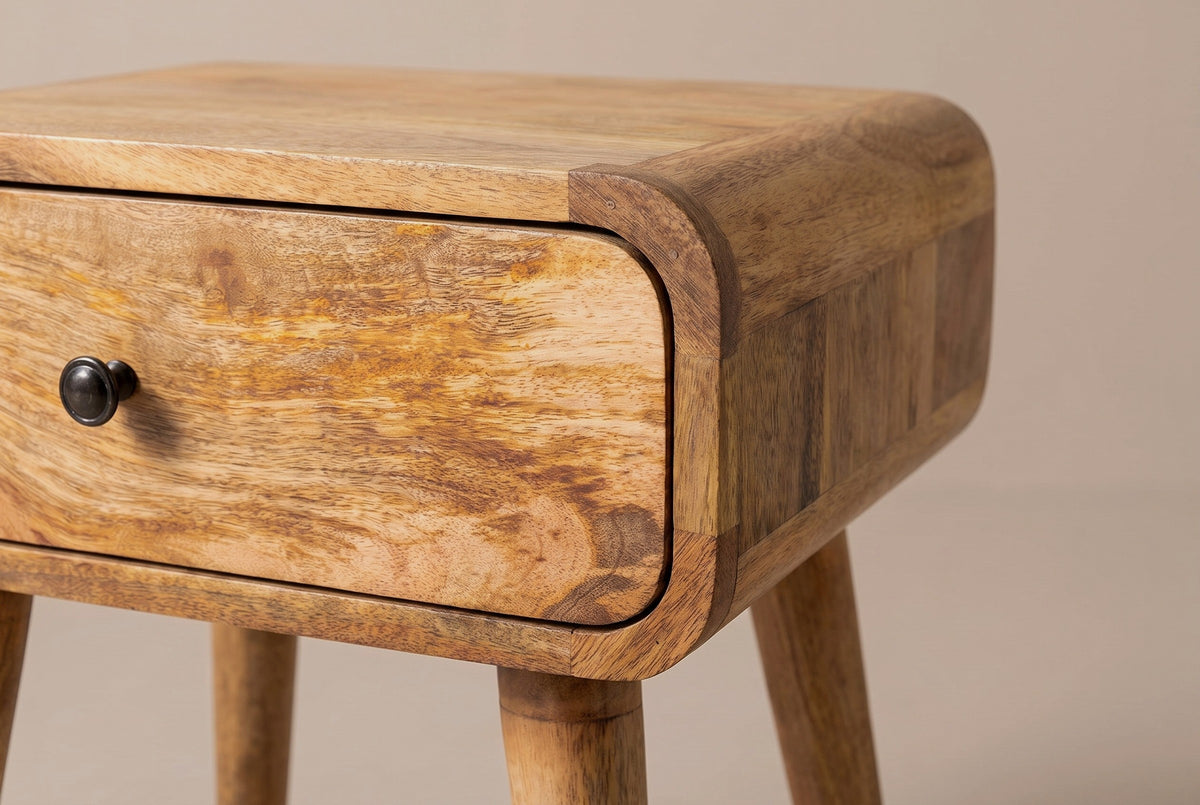 The front corner of the Bonorva nightstand: the top rolling over a rounded corner of curved mango blocks, the flush drawer with its dark round knob and the top of a tapered leg
