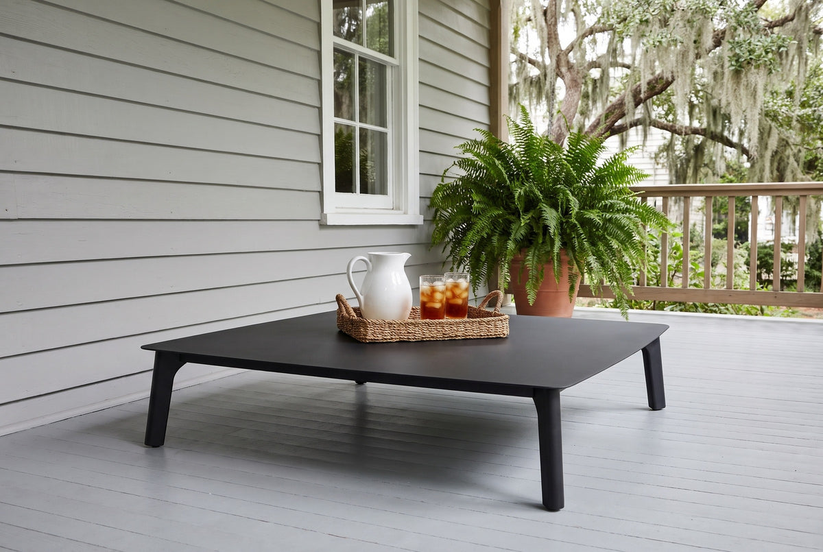 Brucoli black aluminum coffee table on a grey-painted porch floor in Beaufort, South Carolina, grey siding, a fern and live oaks behind (tray, pitcher and plant not included)