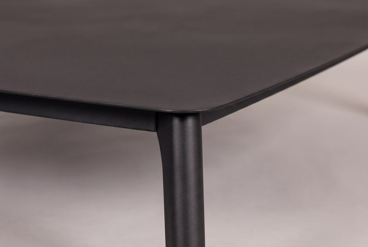 Close-up of the Brucoli table: the rounded corner of the solid aluminum top, the rail and the top of a tapered leg in matte black
