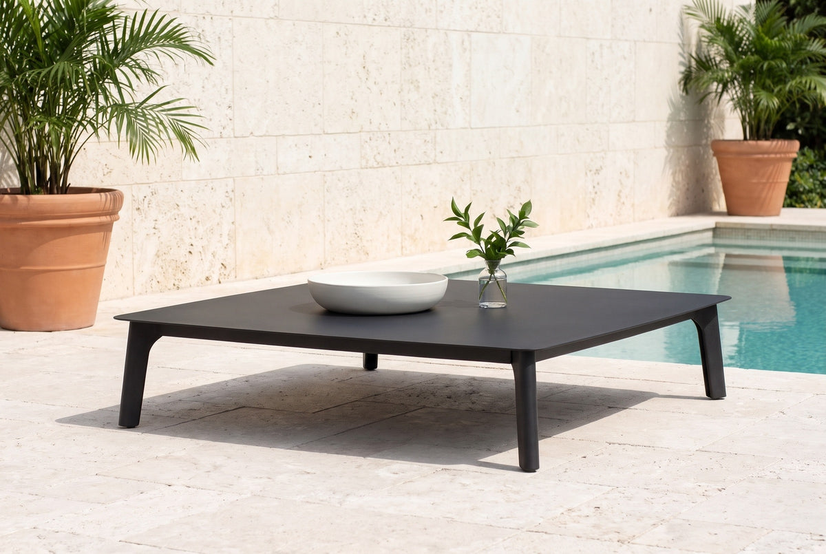 Brucoli black aluminum coffee table on light shellstone pavers by a pool in Naples, Florida, a keystone coral-stone wall and potted palms behind (bowl and vase not included)