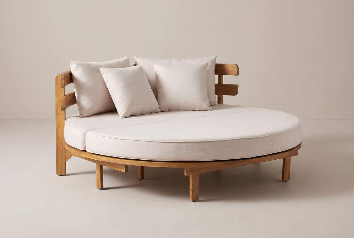 The Butera round acacia daybed with a light beige cushion and four pillows, three-quarter view on a light studio backdrop