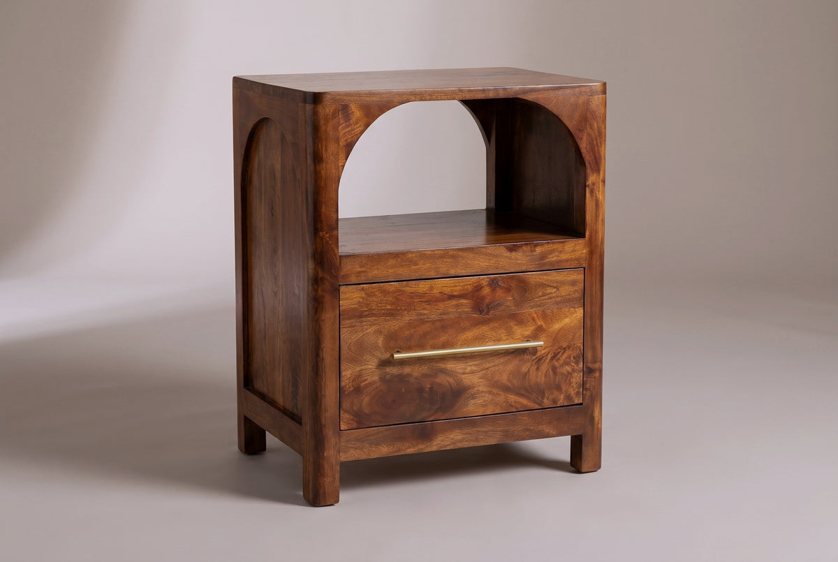The Cabras nightstand in chestnut mango with an open arched cubby, one drawer with a brass bar pull and four short square legs, three-quarter view on a light studio backdrop