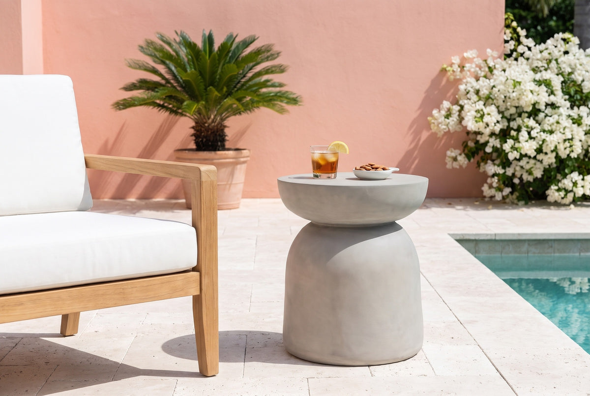 Cammarata light grey concrete dish-top bell side table with a glass of iced tea and a dish of almonds beside a teak armchair on a travertine pool terrace in Naples, Florida, a coral-pink stucco wall and white bougainvillea behind (chair and decor not included)