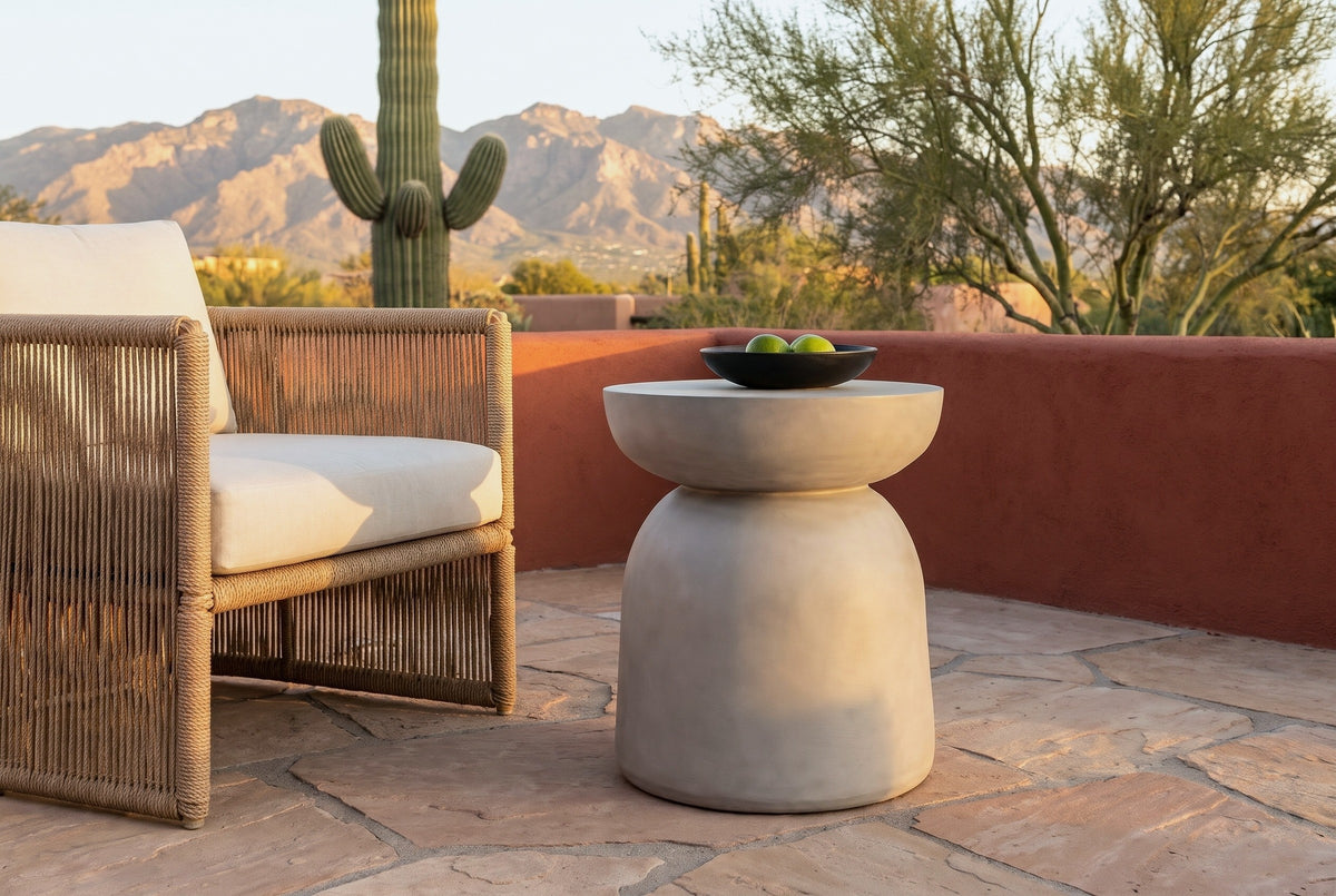 Cammarata light grey concrete dish-top bell side table with a black bowl of limes beside a woven rope armchair on a sandstone patio in Tucson, Arizona at golden hour, a terracotta-red plaster wall, a saguaro and the Santa Catalina mountains behind (chair and decor not included)