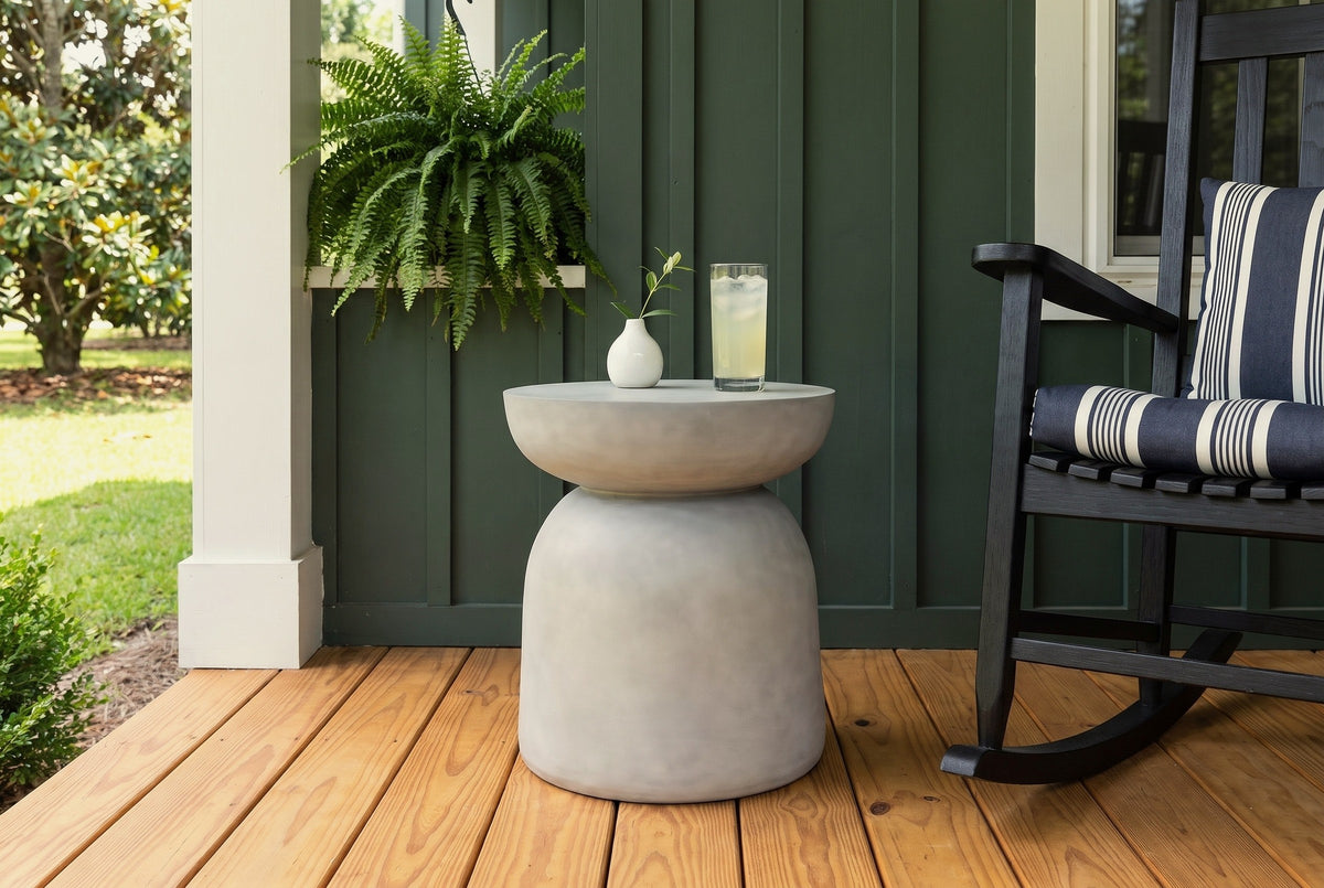 Cammarata light grey concrete dish-top bell side table with a glass of lemonade and a white bud vase beside a black rocking chair on a heart pine porch in Wilmington, North Carolina, a forest-green board-and-batten wall behind (chair and decor not included)