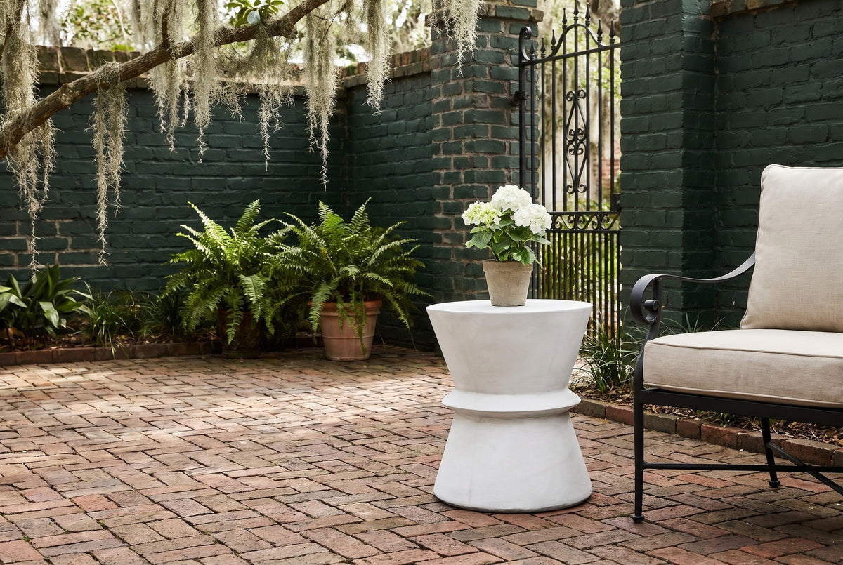 Capizzi concrete side table on herringbone brick in a Savannah courtyard with a green painted brick wall and ferns, beside a wrought-iron armchair (chair and plant not included)