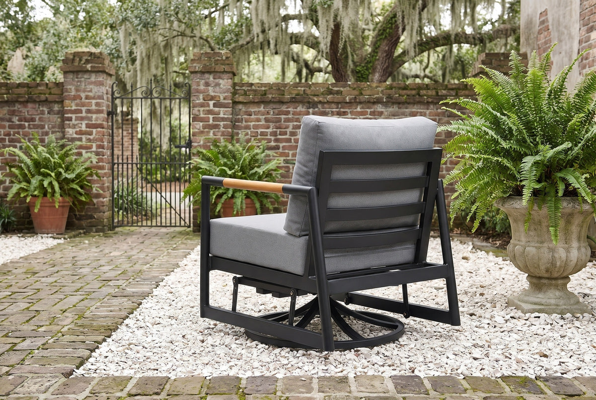 The Carini swivel glider lounge chair seen from behind in a walled Savannah garden courtyard: black aluminum slatted back, teak arm and gray cushions on crushed oyster shell, old brick walls, an iron gate and live oaks with Spanish moss beyond
