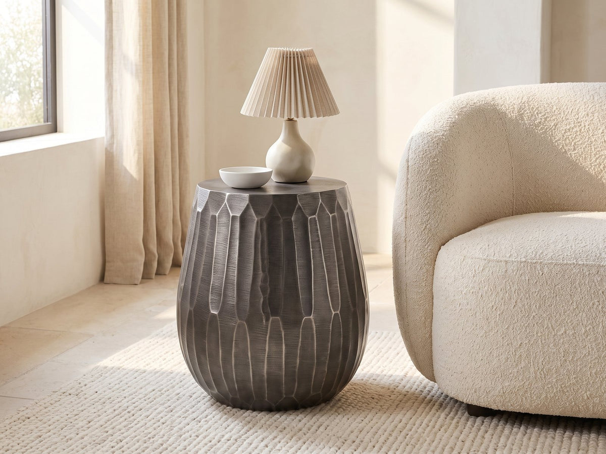 Chianciano Cast Aluminum Faceted Drum Side Table with a lamp and a ceramic bowl beside a cream boucle armchair in a bright plaster room