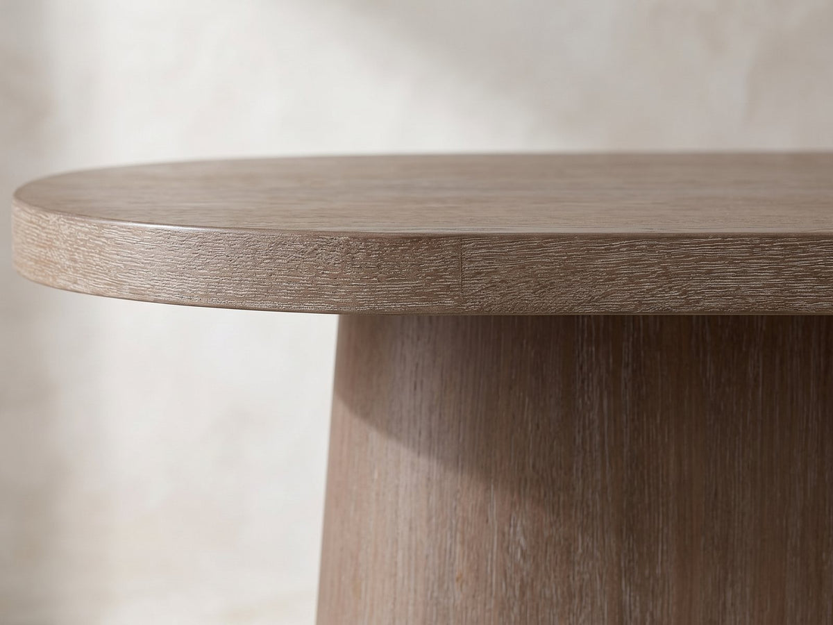 Close-up of the Cotronei 84-Inch Oval Cocoa Wood Veneer Pedestal Dining Table: the square edge of the oval top over the flared slab pedestal