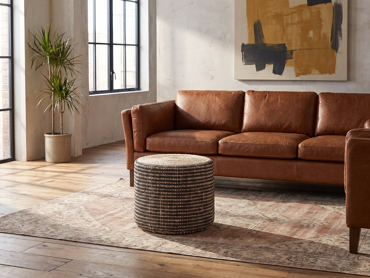Dolceacqua Natural Braided Jute Round Pouf on a vintage rug in front of a leather sofa in an industrial loft