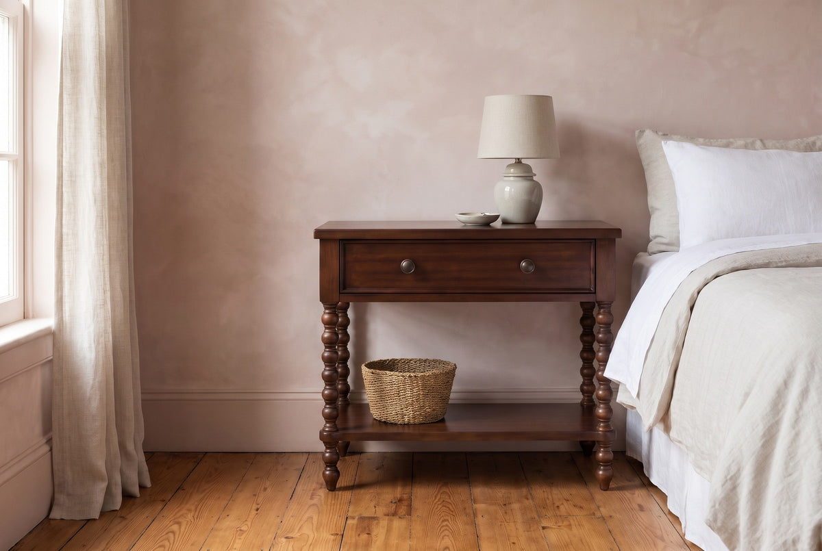 Dorgali Morocco Brown spindle-leg nightstand with a ceramic lamp and a seagrass basket on the shelf beside a bed in a Charleston bedroom with a blush lime-wash plaster wall and a heart-pine floor