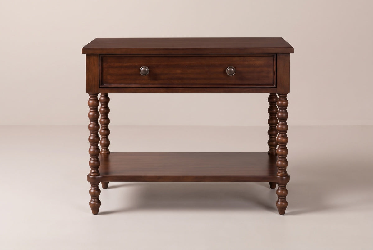 The Dorgali Morocco Brown nightstand, front view on a warm greige studio backdrop: a wide top, one drawer with two knobs, four turned bobbin legs and an open shelf