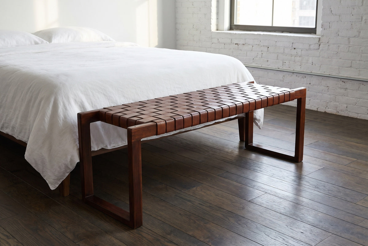 Fasano woven leather bench at the foot of a bed with white linen in a Brooklyn loft bedroom with a white-painted brick wall and dark oak floorboards