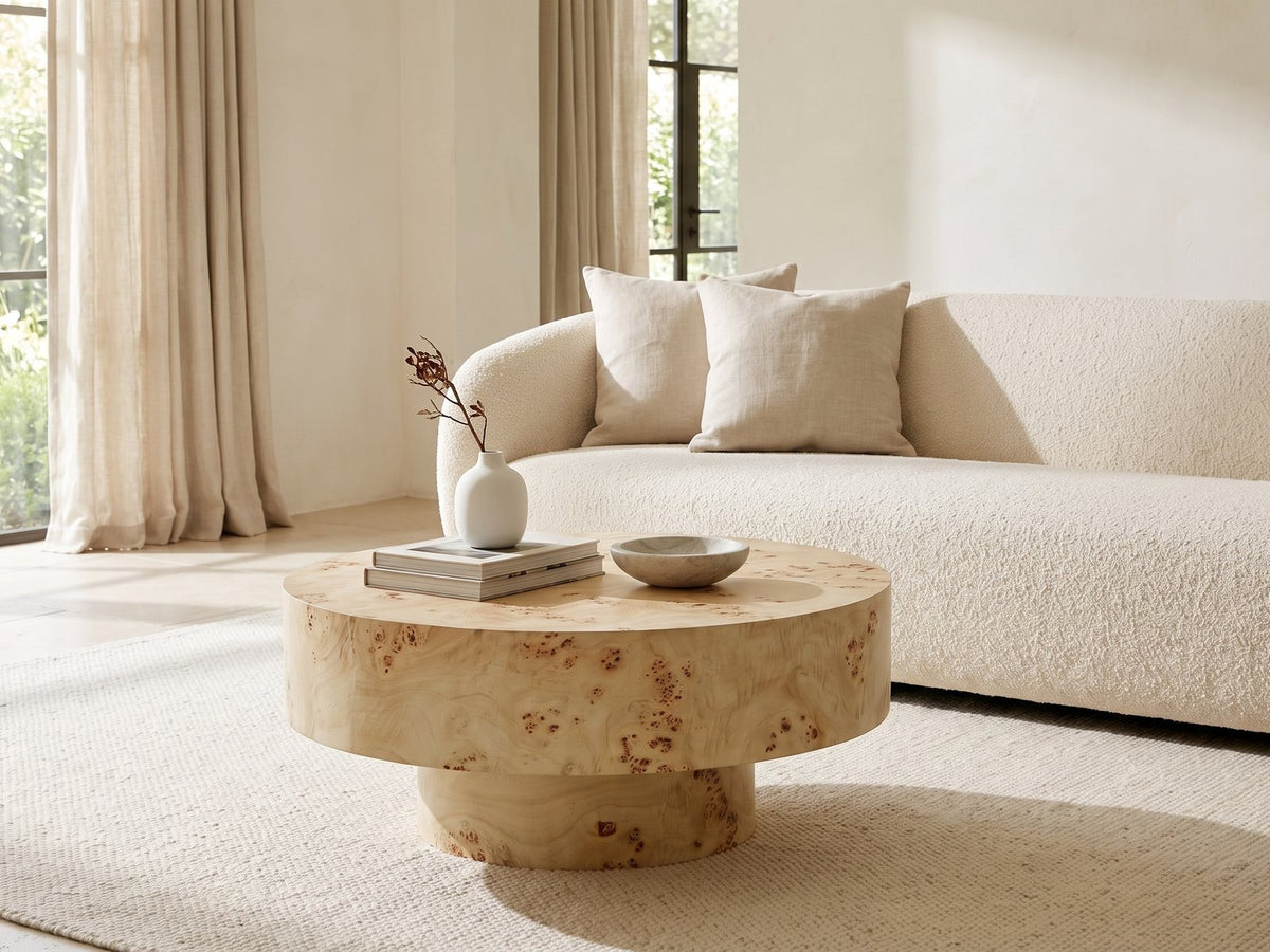 Fossano 31.5-Inch Round Burl Wood Veneer Pedestal Coffee Table styled with books, a vase and a stone bowl in front of a cream sofa in a bright plaster room