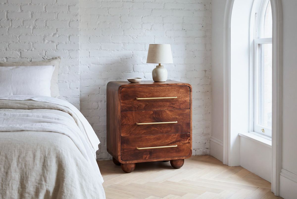 Galtelli chestnut mango nightstand with a ceramic lamp against a whitewashed brick wall in a Brooklyn bedroom with an arched window and a herringbone floor