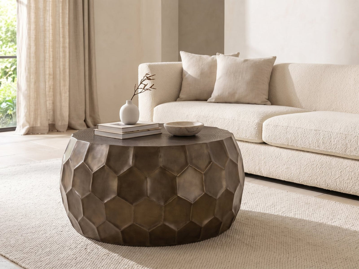 Gavi 32-Inch Round Antique Bronze Faceted Aluminum Drum Coffee Table styled with books, a vase and a stone bowl in front of a cream sofa in a bright plaster room
