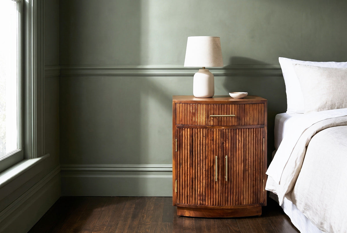 Gavoi fluted mango bow-front nightstand with a ceramic lamp beside a bed in a San Francisco bedroom with a sage green plaster wall and dark oak floorboards