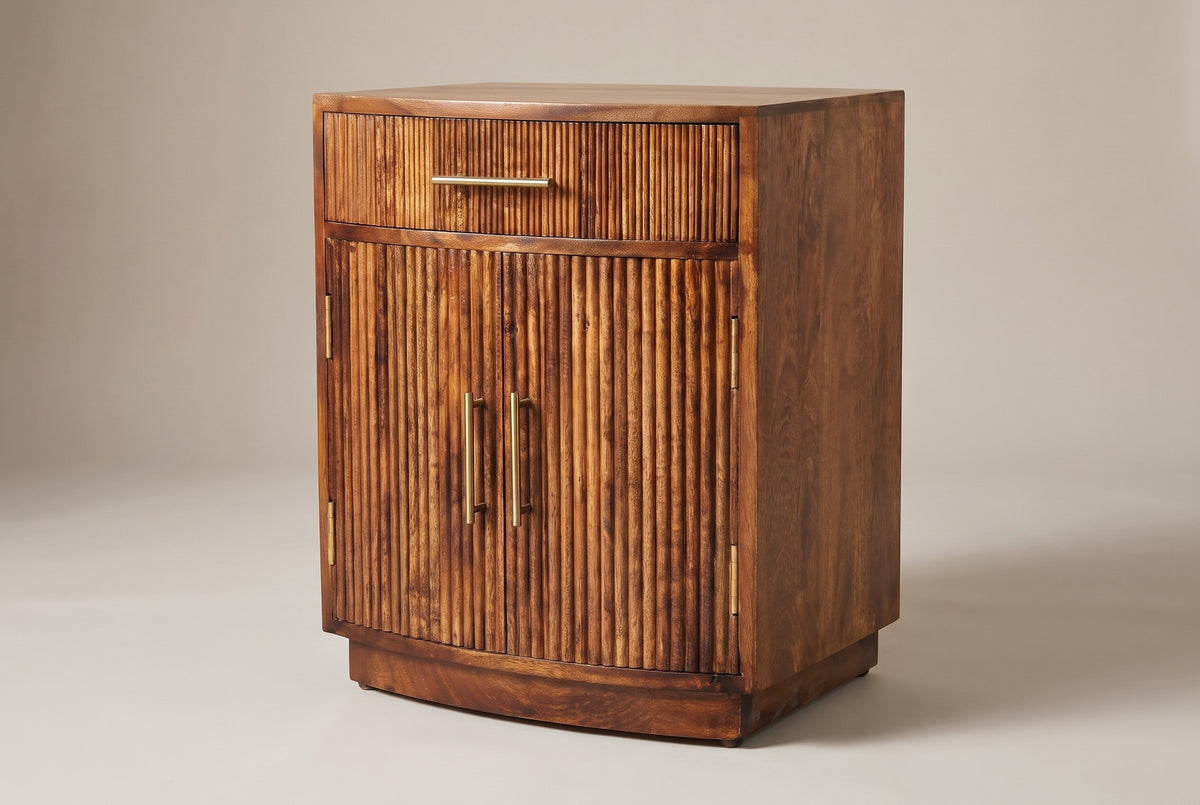 The Gavoi nightstand in mocha mango with a bowed front, one reeded drawer with a brass bar pull and two reeded doors with vertical brass pulls, three-quarter view on a light studio backdrop