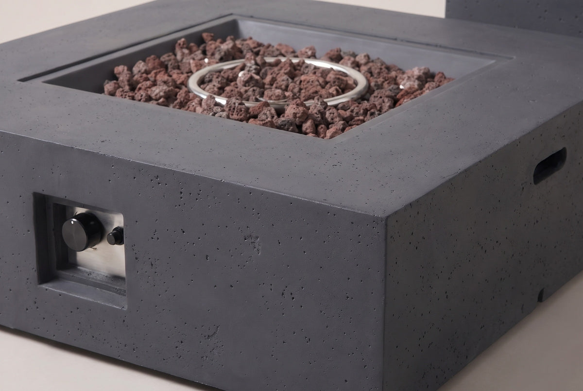 Close-up of the Gela fire pit: the flat dark gray concrete frame with its fine pores, the stepped edge of the square pan, lava rocks and the ring burner
