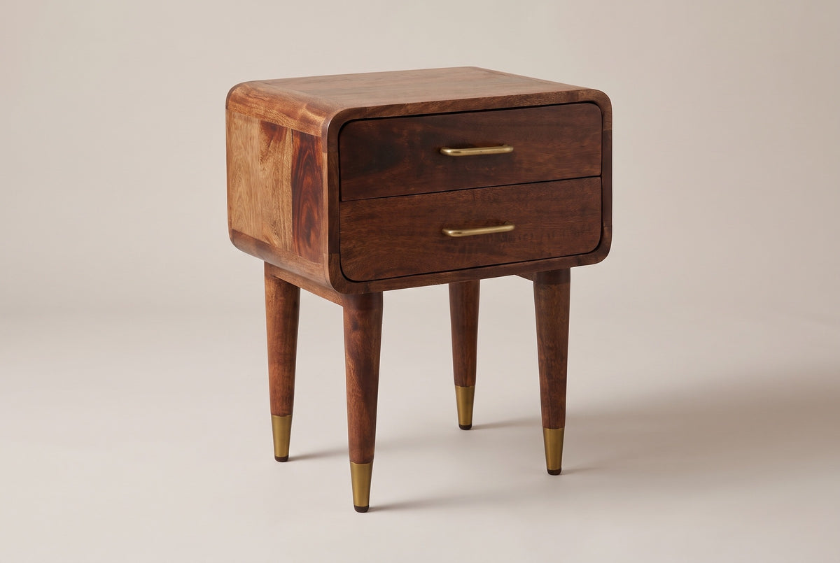 The Ghilarza two-drawer nightstand in chestnut mango with slim brass pulls, rounded corners and four tapered legs with brass caps, three-quarter view on a light studio backdrop