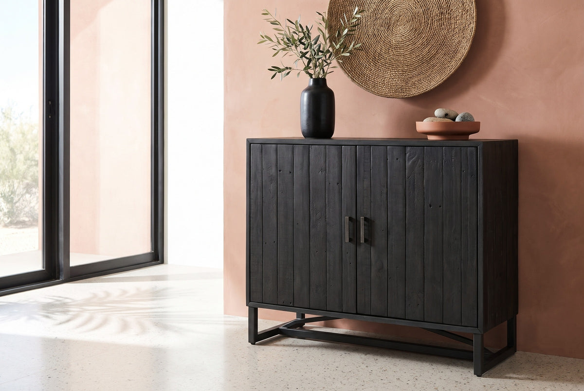 Gravina black reclaimed pine two-door cabinet with an olive branch in a black vase and a woven seagrass disc above it, against a terracotta clay wall in a Scottsdale living room