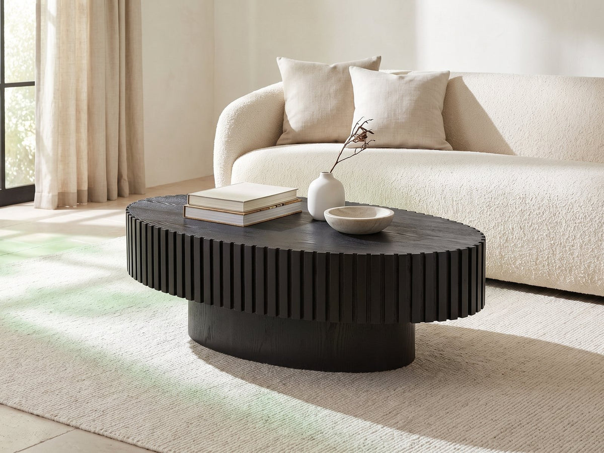 Imola Black Ash Oval Pedestal Coffee Table styled with books, a vase and a stone bowl in front of a cream sofa in a bright plaster room
