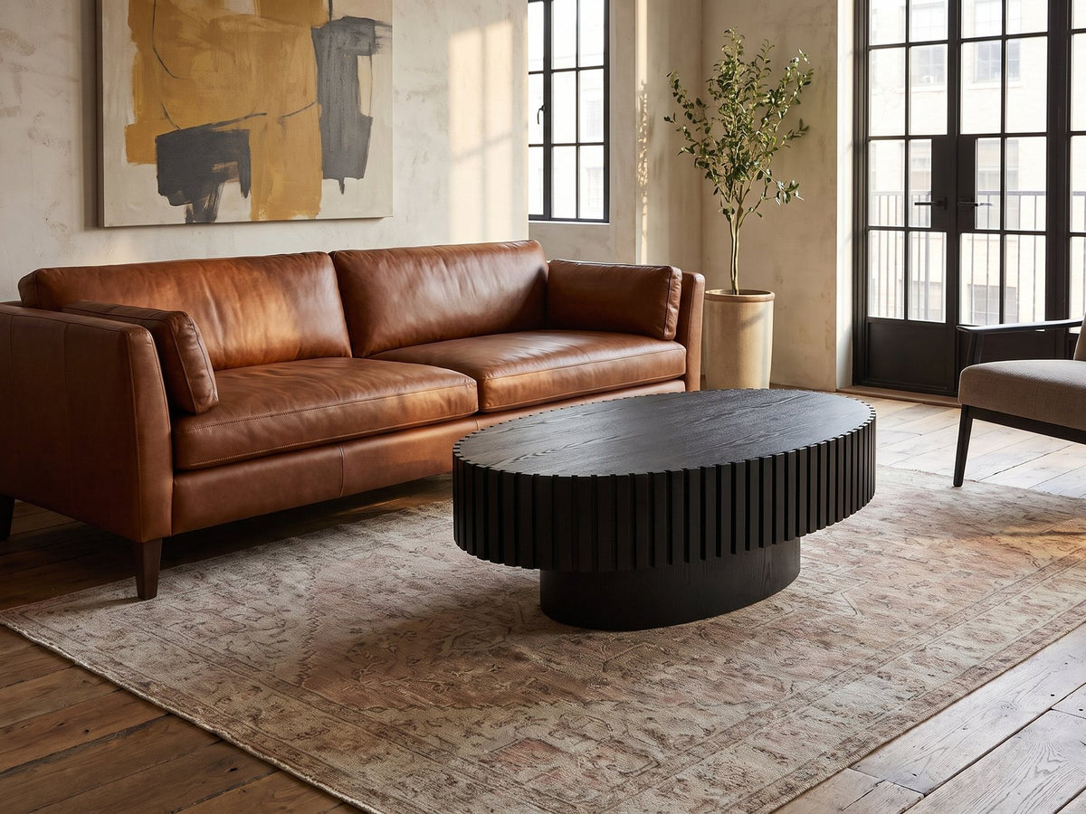 Imola Black Ash Oval Pedestal Coffee Table on a vintage rug in front of a leather sofa in an industrial loft