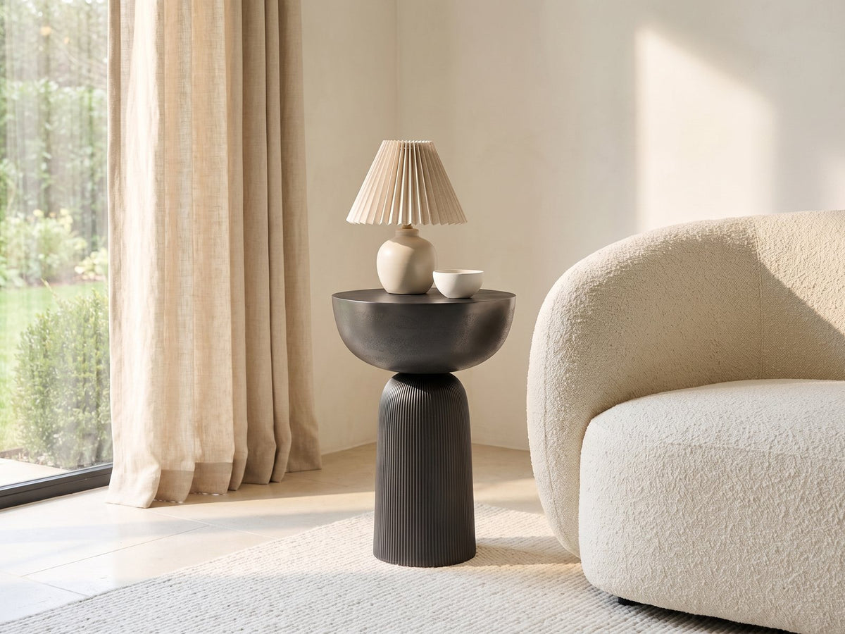 Irsina Cast Aluminum Tulip Pedestal Side Table with a lamp and a ceramic bowl beside a cream boucle armchair in a bright plaster room