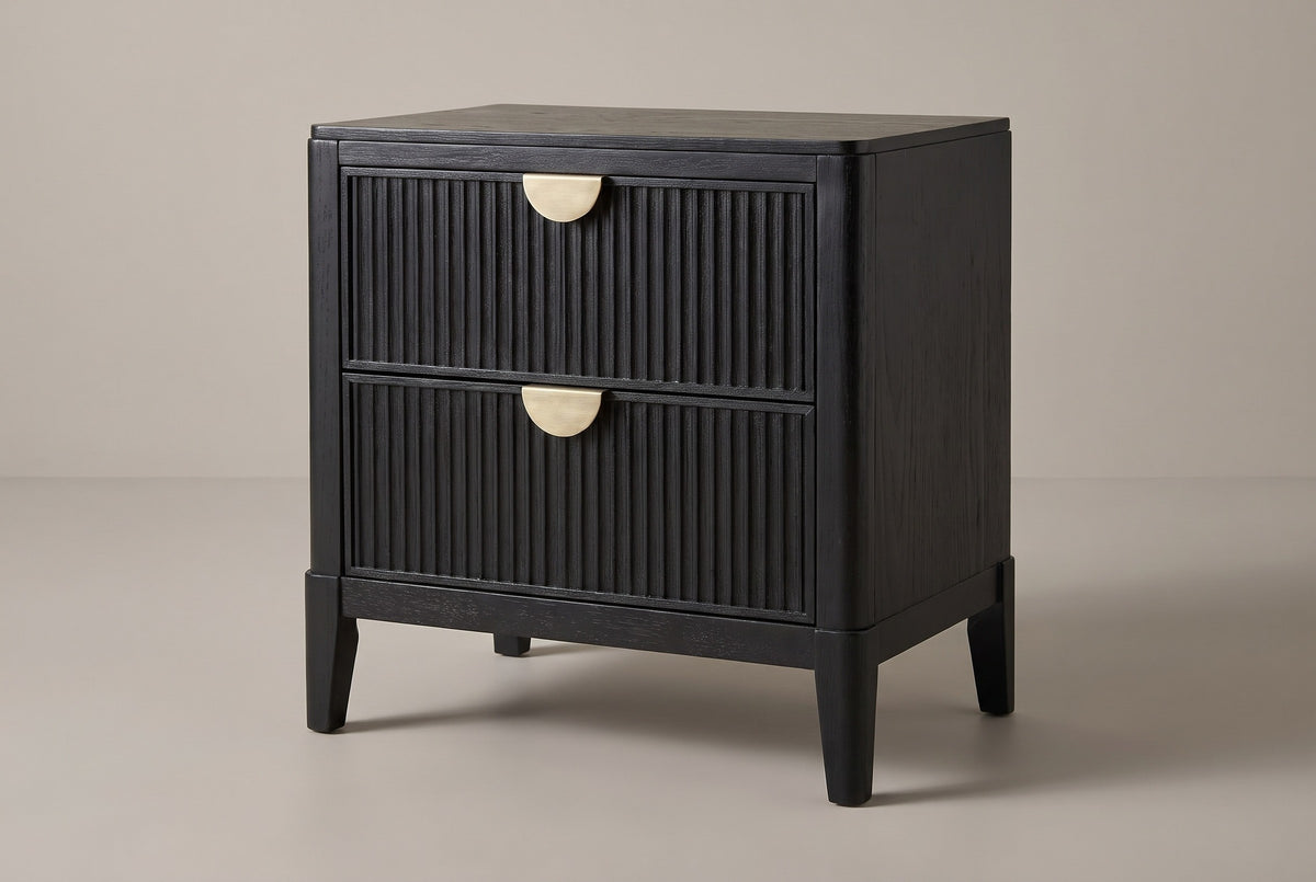 The Isili nightstand in wire-brushed black wood with two fluted drawers, brass half-moon pulls and four square tapered legs, three-quarter view on a light studio backdrop