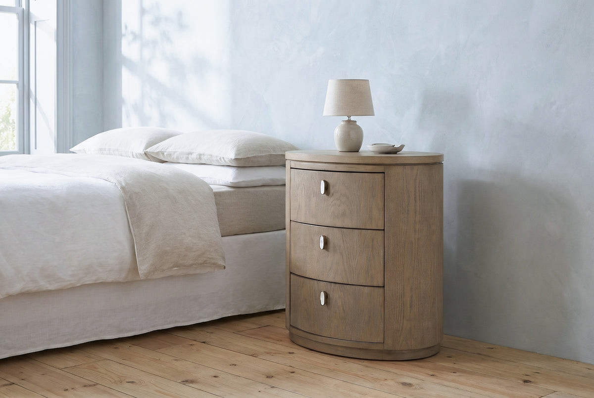 Jerzu cerused toffee oval nightstand with a ceramic lamp beside a bed in a Charleston bedroom with a pale blue-grey plaster wall and a pine floor