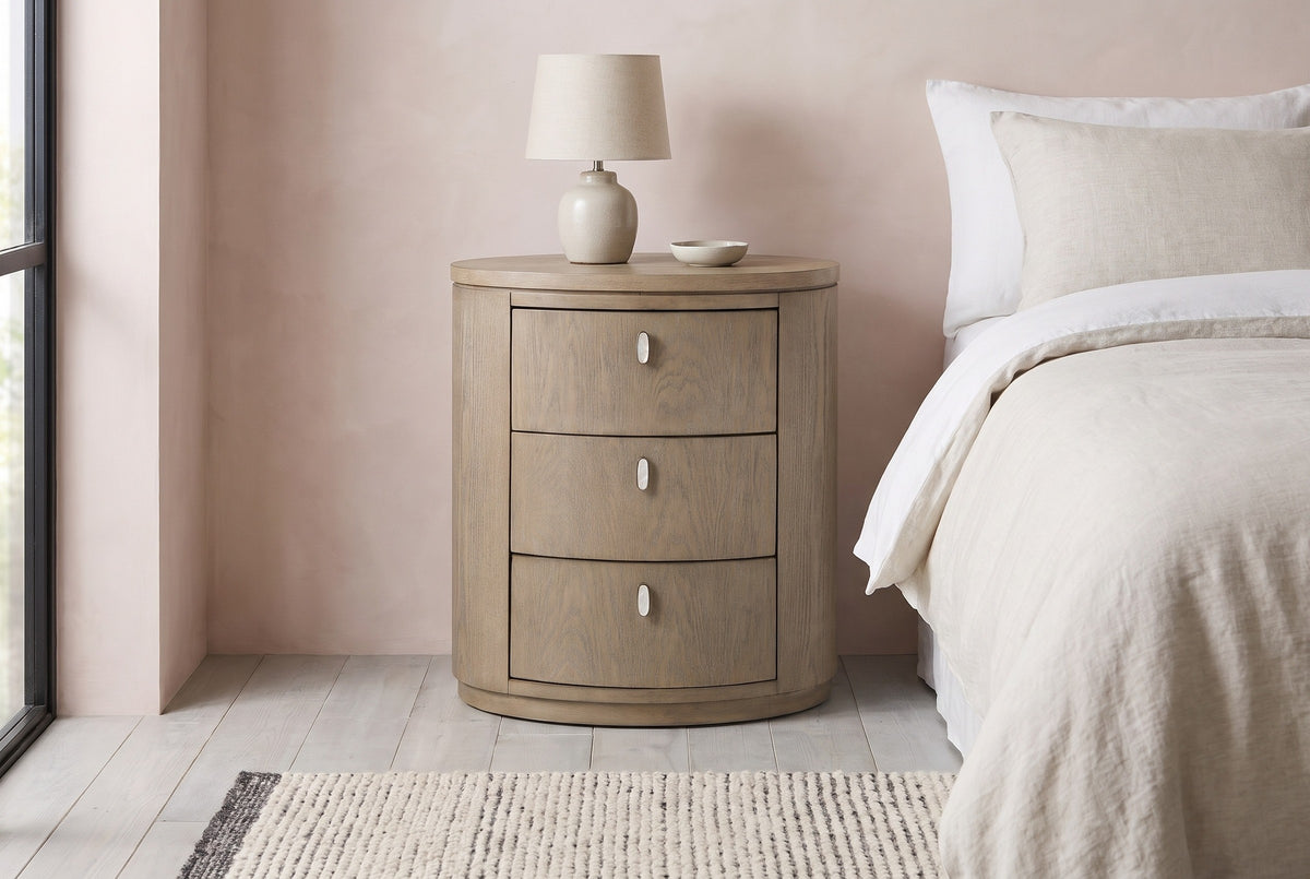 Jerzu cerused toffee oval nightstand with a ceramic lamp against a blush pink limewash wall in a Denver bedroom with grey-washed boards and a black steel window