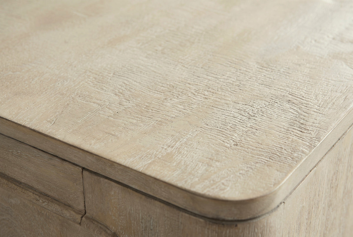 Close-up of the weathered mango on the Laconi nightstand: a pale sandy wood with fine rippled grain and a light white wash, at the rounded corner of the top