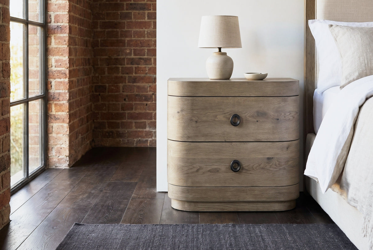 Lanusei knotty oak nightstand with a ceramic lamp beside an upholstered bed in a Chicago loft bedroom with exposed brick, a steel-framed window and a dark wood floor