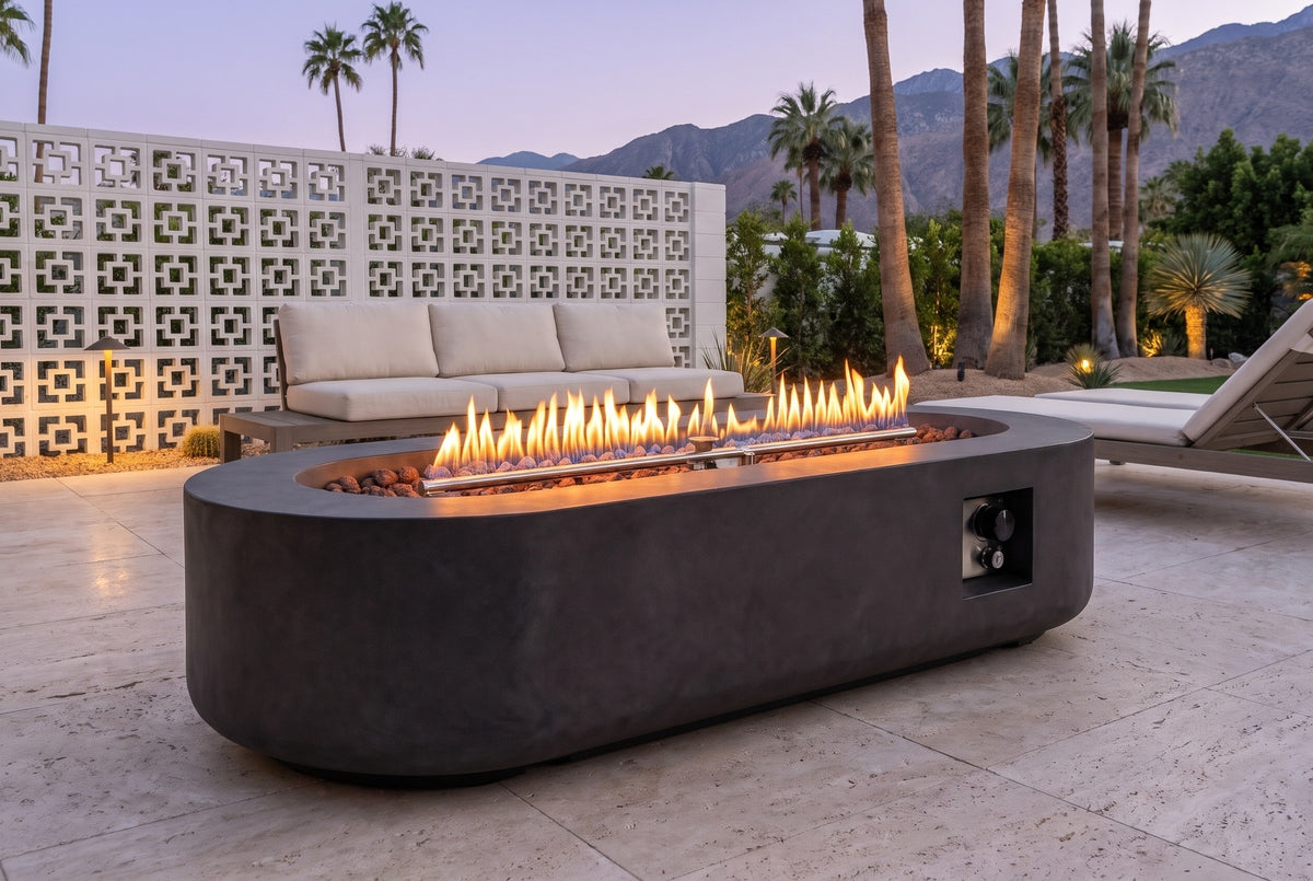 Licata oval concrete fire pit table lit at blue hour on travertine pavers in a Palm Springs patio, a white breeze-block wall, palms and mountains behind (sofa and lounge chair not included)