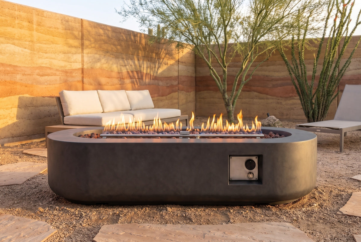 Licata oval concrete fire pit table lit at golden hour on decomposed granite in a Tucson desert patio, a layered rammed-earth wall, a palo verde tree and an ocotillo behind (sofa and lounge chair not included)