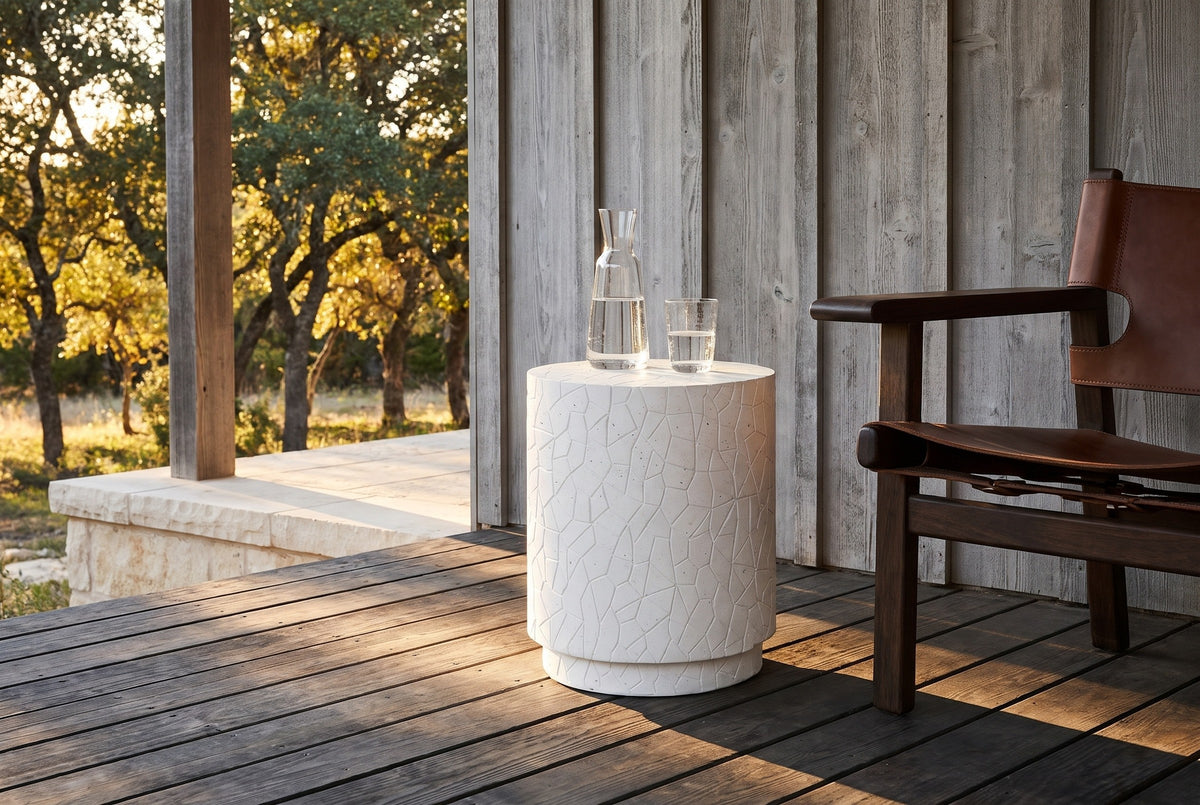 Linguaglossa white concrete drum side table on a dark plank porch in the Texas Hill Country, a weathered cedar wall and live oaks behind, a carafe and glass on top (chair and glassware not included)