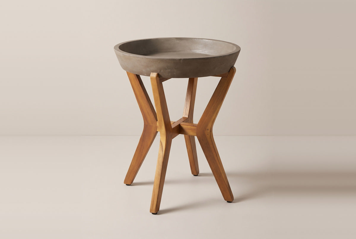 The Linosa side table: a round concrete tray top with a raised rim on four chevron-shaped acacia legs joined by a cross stretcher